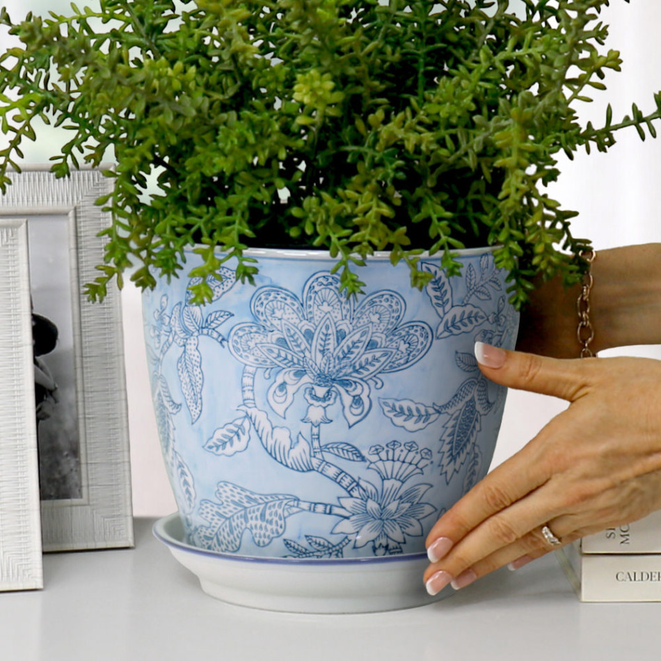 Hands positioning a blue floral ceramic plant pot and saucer with fine green foliage beside silver frames