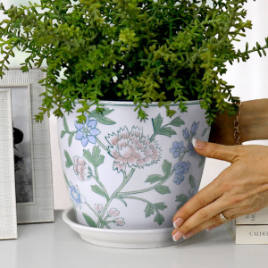 Close-up of hands positioning a white floral ceramic plant pot with green foliage and a matching saucer