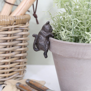 Cast iron kitten hanging from a pale plant pot with green foliage, beside a woven basket and garden tools