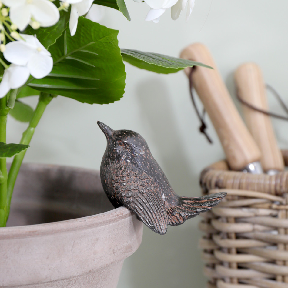 Vintage brown bird pot hanger on a pale planter with white flowers, beside a woven basket holding garden tools