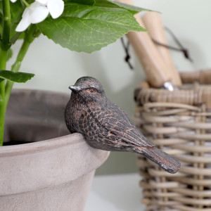 Brown resin bird perched on a pale plant pot beneath green leaves, beside a woven basket holding garden tools