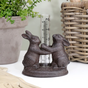 Bunny rabbit rain gauge on a white bench beside a potted plant, woven basket and gardening gloves