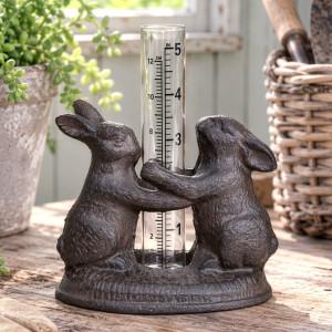 Cast iron bunny rabbit rain gauge with glass measuring tube on a timber bench beside a plant and woven basket