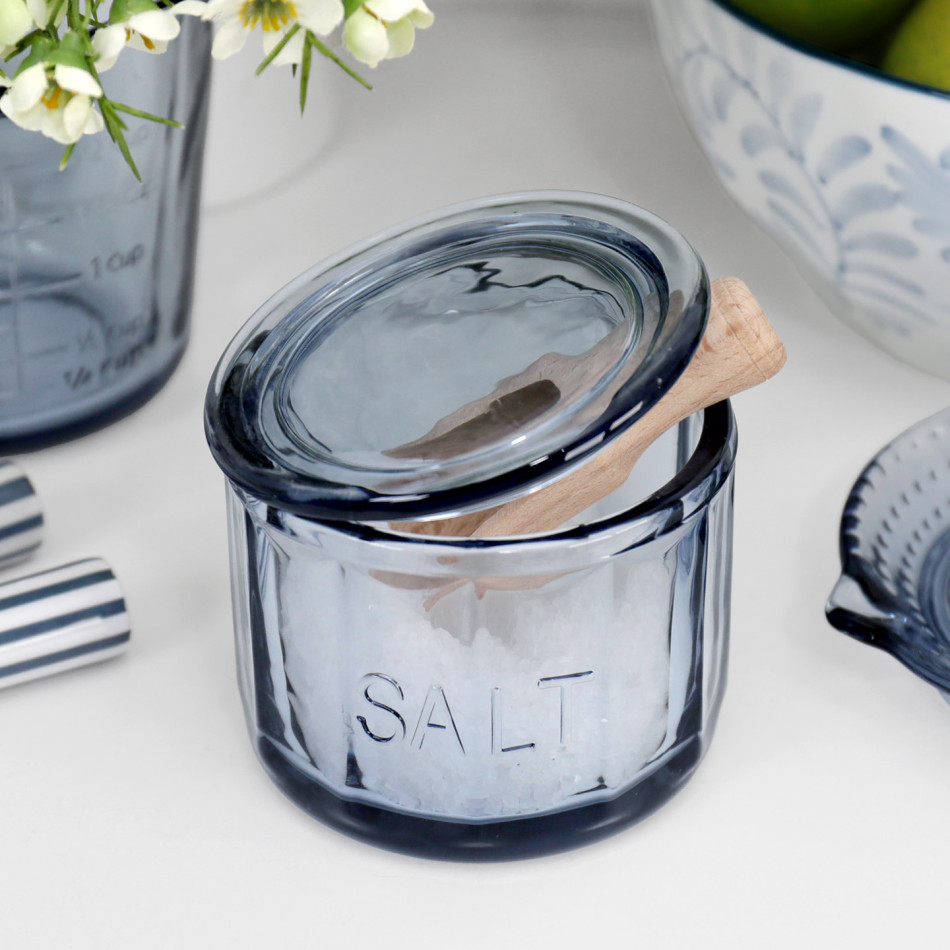 Soft blue glass salt pot with raised SALT lettering, glass lid and wooden scoop on a white kitchen bench
