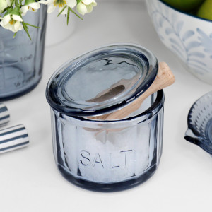 Soft blue glass salt pot with raised SALT lettering, glass lid and wooden scoop on a white kitchen bench