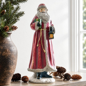 Father Christmas in a distressed red coat holding a lantern and green gift, styled with pine cones beside a window