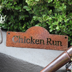 Rusty steel Chicken Run sign with cutout lettering and two fixing holes, styled against greenery beside garden tools