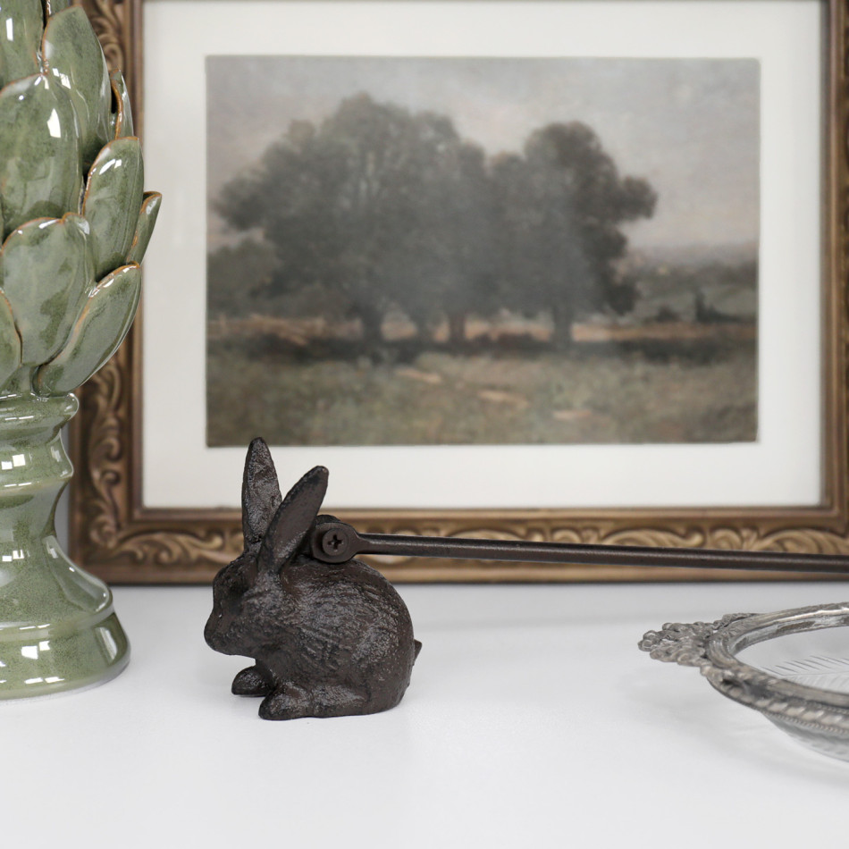 Cast iron rabbit candle snuffer styled on a white console beneath framed country artwork.