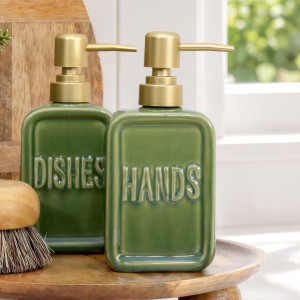 Green ceramic 'Hands' soap dispenser with gold-toned pump beside matching 'Dishes' dispenser on a wooden tray