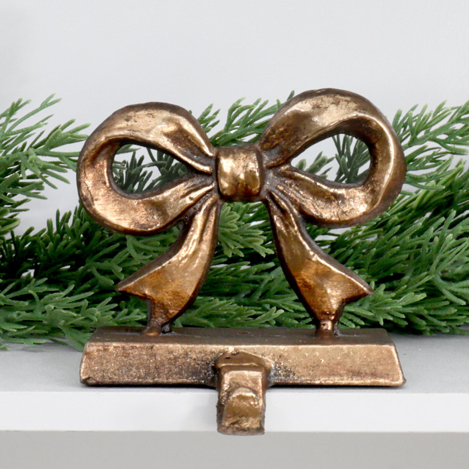 Clara aged gold bow stocking hook on a white shelf with green foliage behind.