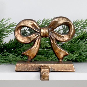 Clara aged gold bow stocking hook on a white shelf with green foliage behind.