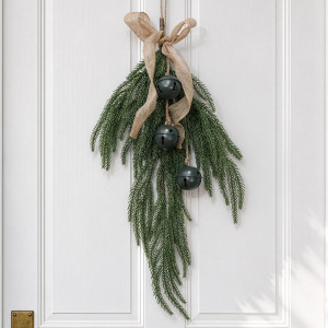 Glad Tidings Christmas swag with cascading green foliage, three dark green metal bells and a hessian bow against white panelling.