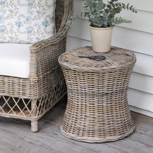 Antique-grey rattan side table with a plant beside a floral-cushioned cane chair