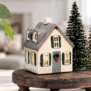 Cream ceramic Christmas house with glowing windows and a grey roof beside a green Christmas tree