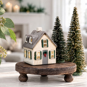 Illuminated ceramic Christmas house on a round wooden stand beside two decorative green Christmas trees