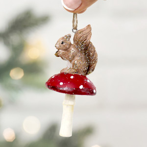 Close-up of glittered squirrel on a red spotted toadstool, held by its hanging loop against blurred Christmas lights