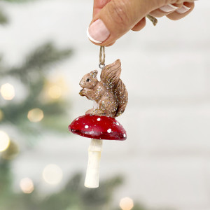 Squirrel on toadstool Christmas decoration held by its twine loop with greenery and warm lights behind