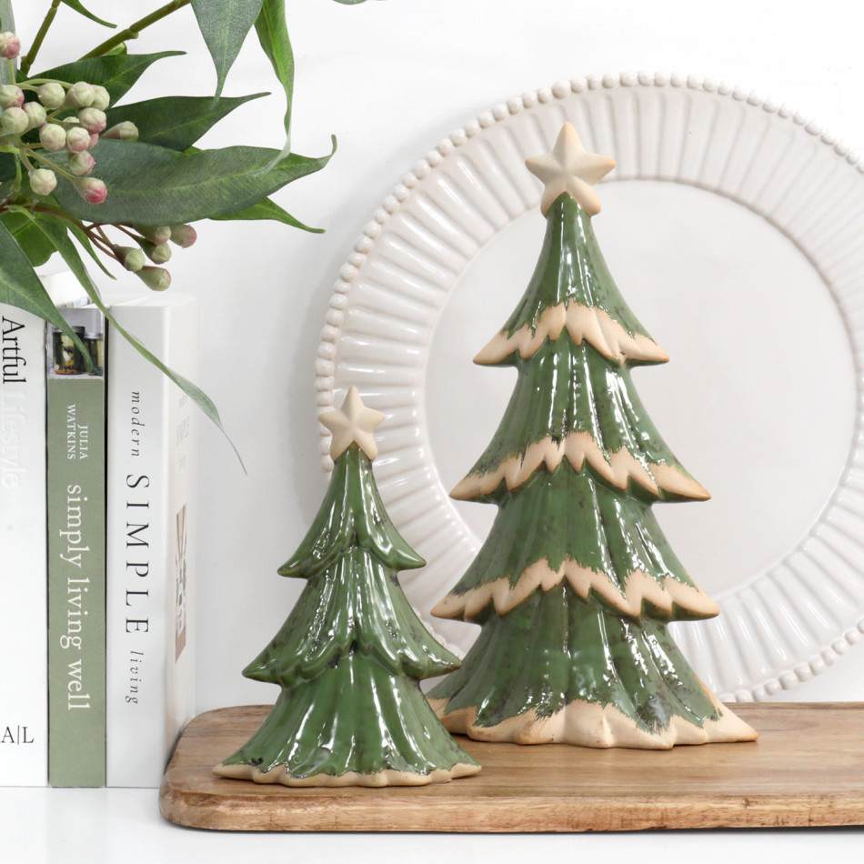 Small and large green ceramic Christmas trees on a wooden board, styled with books, leafy stems and a white decorative plate.