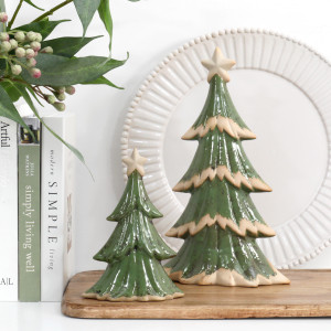 Small and large green ceramic Christmas trees on a wooden board, styled with books, leafy stems and a white decorative plate.