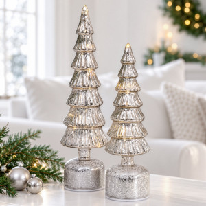 Small silver light-up Christmas tree beside the large size on a white table with greenery and silver baubles