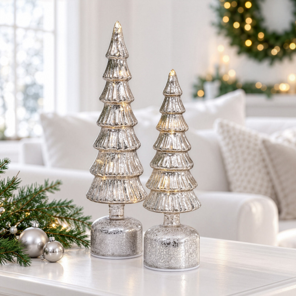 Small and large illuminated silver Christmas trees displayed in a living room beside festive greenery