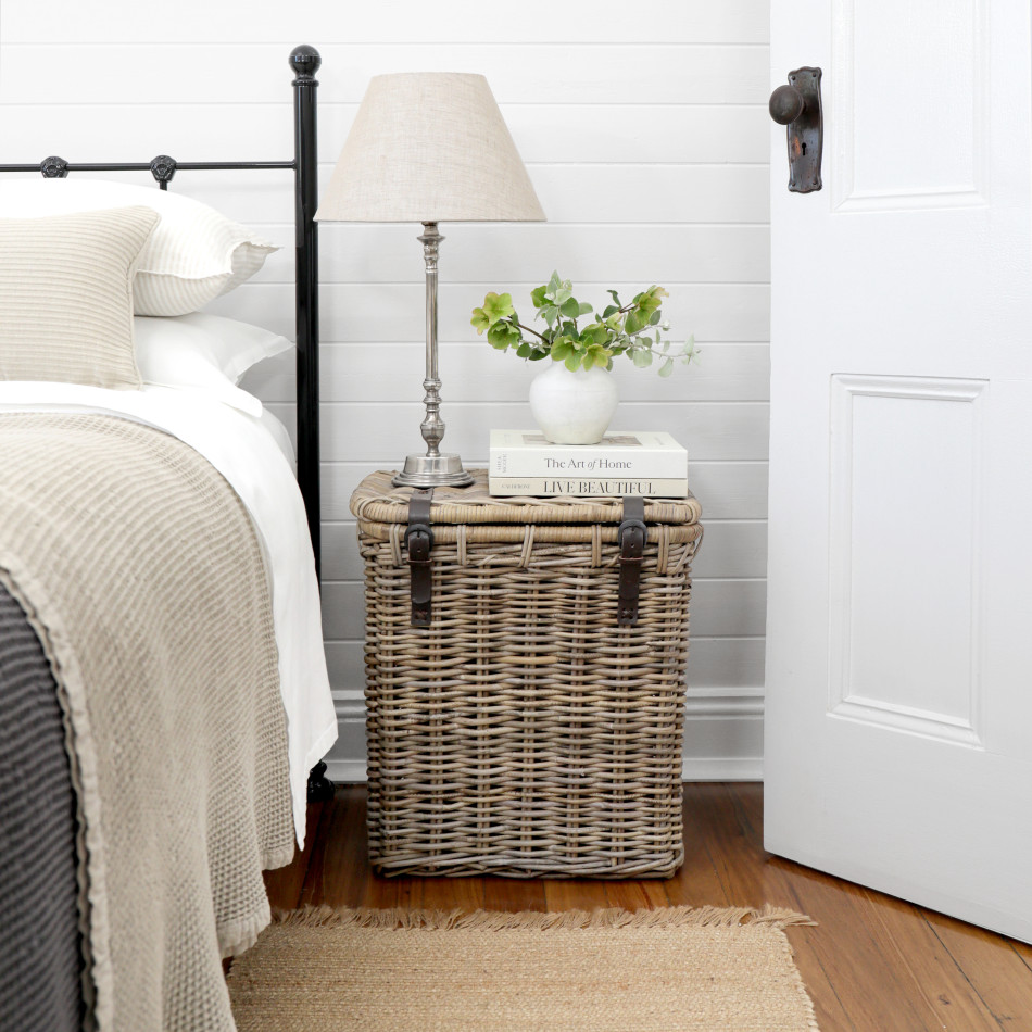 Tall rattan bedside trunk with leather straps in a neutral bedroom setting