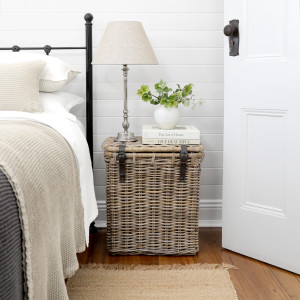 Tall rattan bedside trunk with leather straps in a neutral bedroom setting