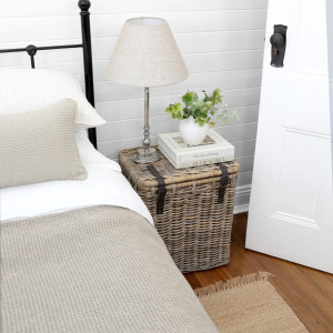 Grey-brown rattan trunk and lamp styled beside a bed with neutral linen