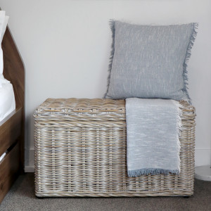 Greywash rattan blanket box beside a bed with a cushion and folded throw on top
