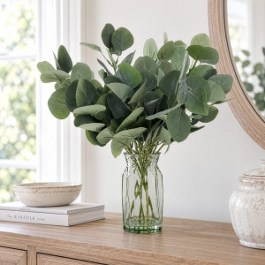 Artificial eucalyptus arrangement in a green-tinted glass vase beneath a round mirror