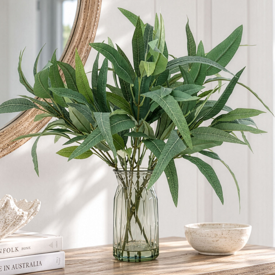 Spear-shaped artificial eucalyptus leaves in a green-tinted glass vase on a wooden console