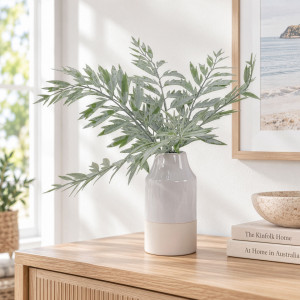 Artificial fern foliage in a tall ceramic vase styled on a light wood sideboard