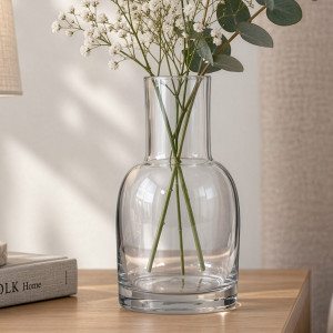 Clear bottle-shaped glass vase holding white flowers and eucalyptus on a wooden table